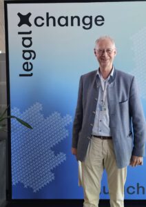 Smiling older man in a blue blazer and beige pants standing in front of a gradient blue display reading 'lead change'.